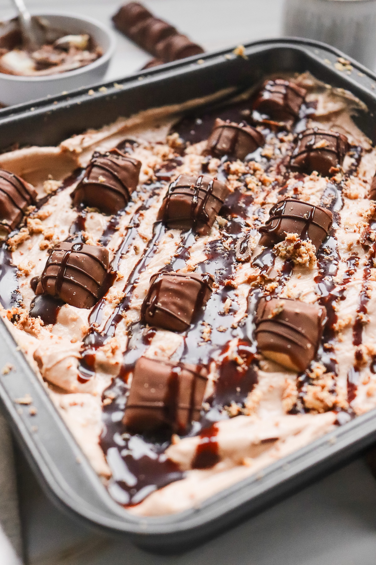 Ice cream topped with chocolate drizzle and chopped chocolate bars in a rectangular baking pan creates the perfect Kinder Bueno No Bake Cake experience.