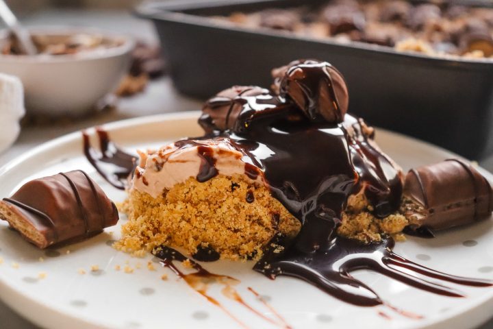 A Kinder Bueno No Bake Cake dessert topped with chocolate sauce and candies, with cookie crumbs on a white plate.