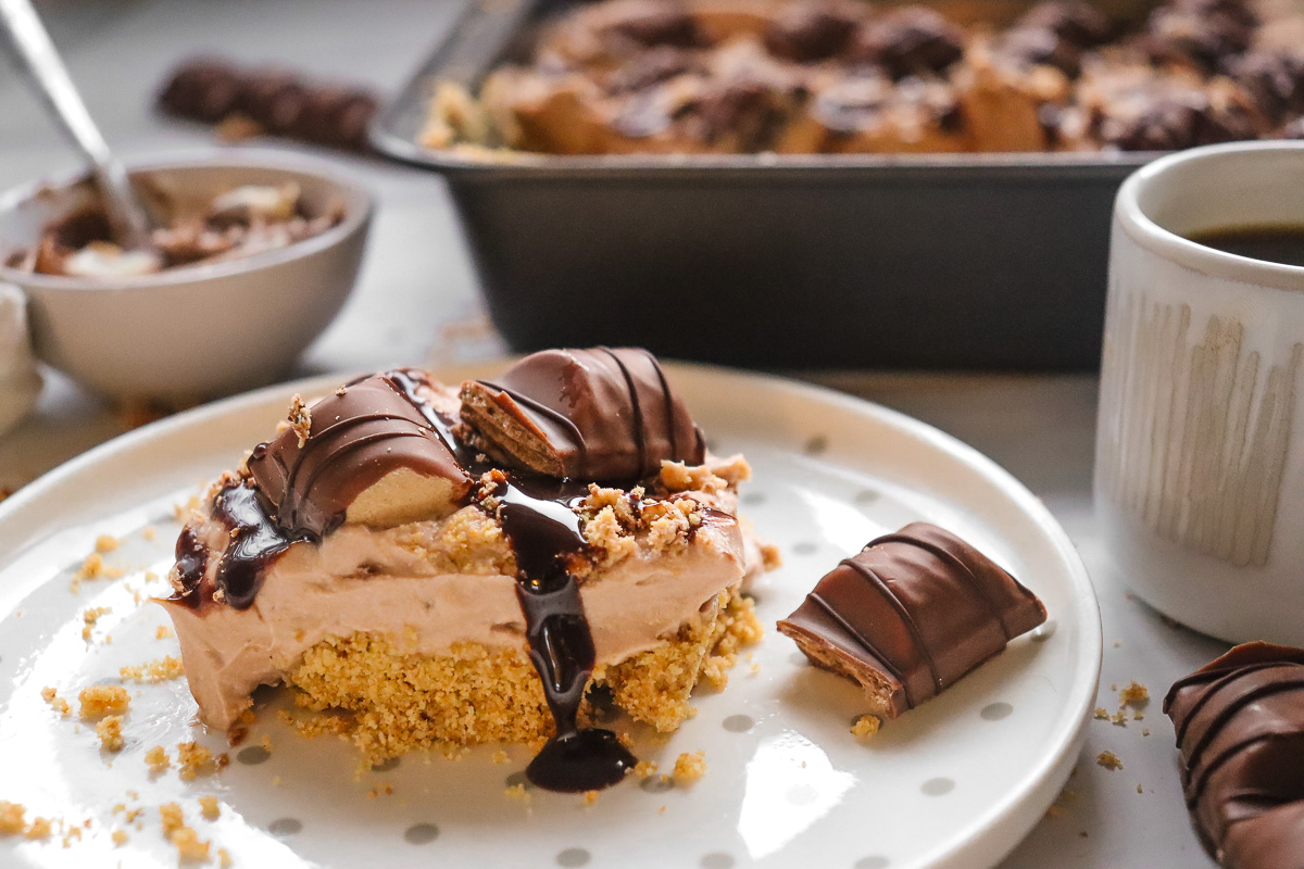 A Kinder Bueno No Bake Cake dessert bar topped with chocolate, cream, and wafer pieces sits on a plate next to a mug and baking pan.