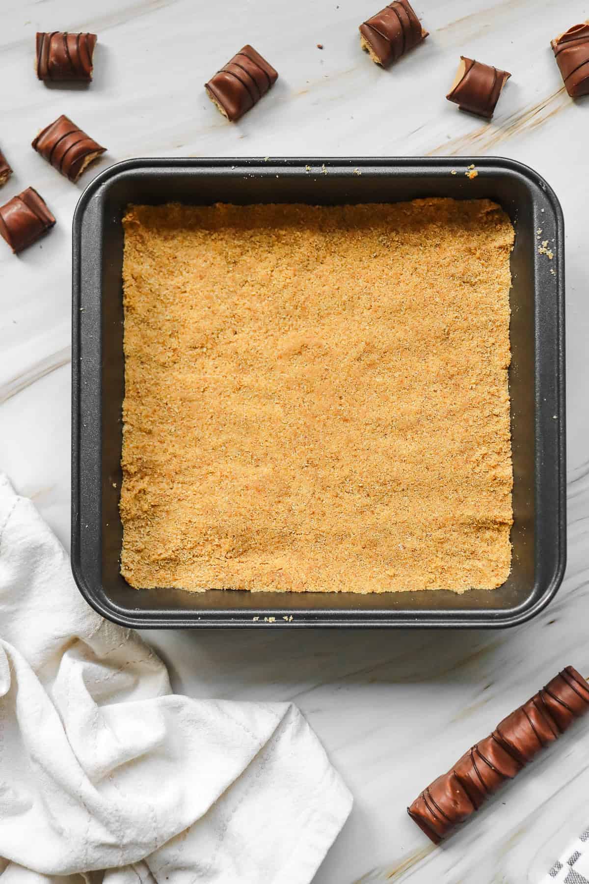 Square baking pan with a graham cracker crust, surrounded by pieces of chocolate candy on a marble surface—perfect for creating a delicious Kinder Bueno No Bake Cake.