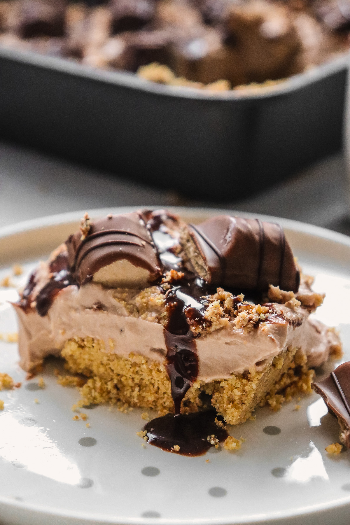 A slice of Kinder Bueno No Bake Cake sits on a white plate, topped with chocolate sauce and candy pieces, creating a creamy chocolate and cream dessert that’s as irresistible as it is decadent.