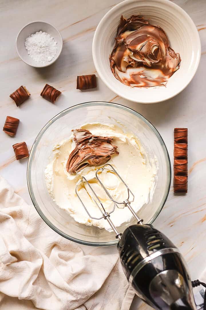 Electric mixer with cream and chocolate in a bowl, with chocolate bars and powdered sugar nearby, perfect for preparing a decadent Kinder Bueno No Bake Cake.