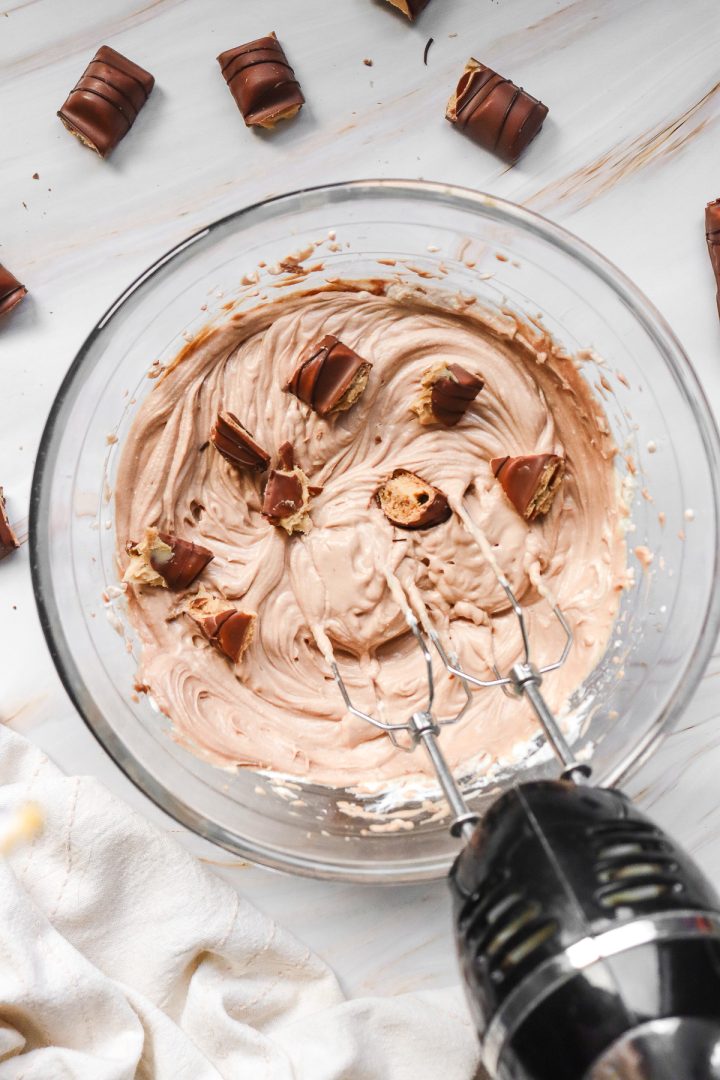 A mixing bowl of chocolate cream with candy pieces for a Kinder Bueno No Bake Cake and a hand mixer on a marble counter.