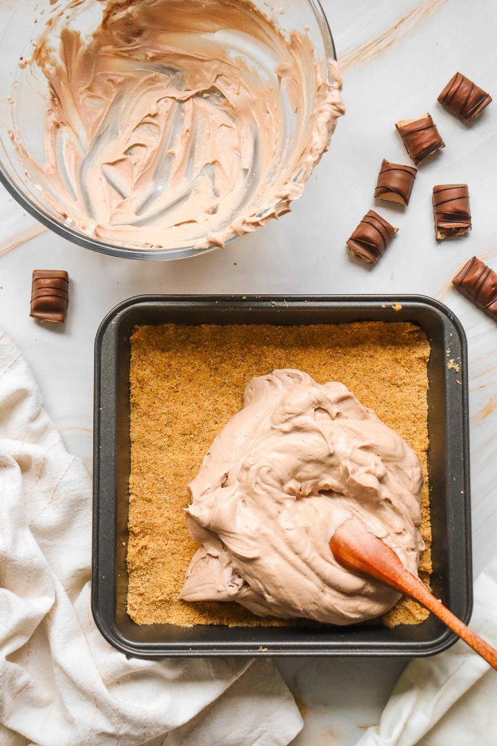 A baking pan with graham crust and chocolate cream, perfect for a Kinder Bueno No Bake Cake, sits beside a bowl with leftovers and chopped chocolate pieces nearby.