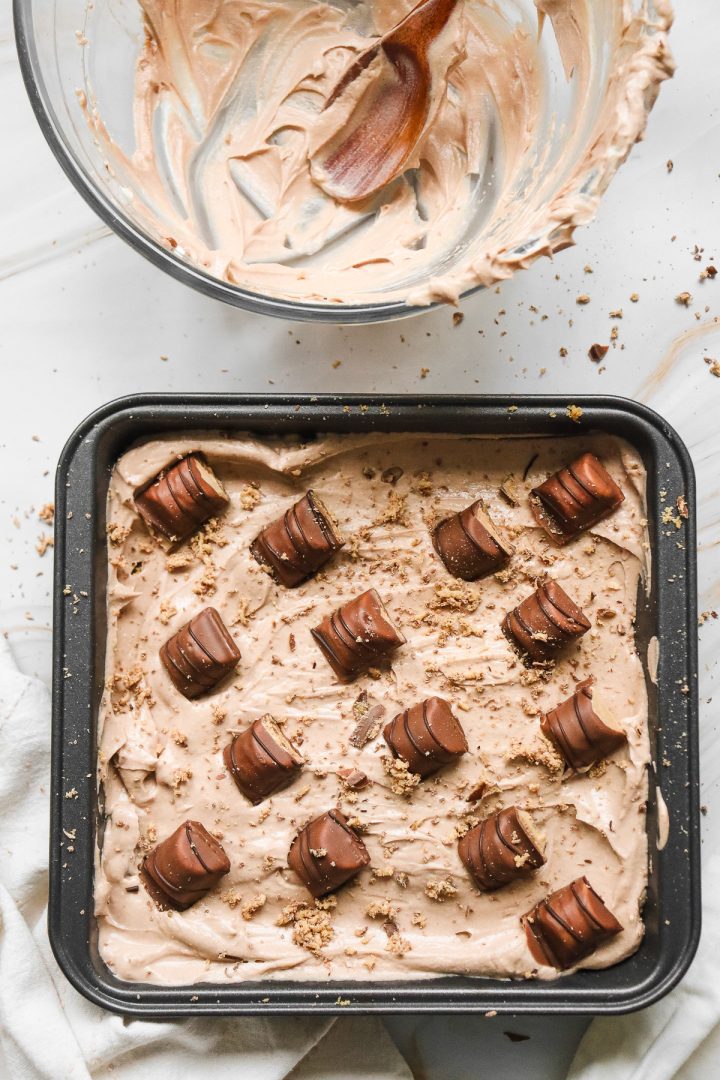 Square pan of rich chocolate mousse topped with chocolate pieces, inspired by a Kinder Bueno No Bake Cake, with a mixing bowl and spoon beside it.