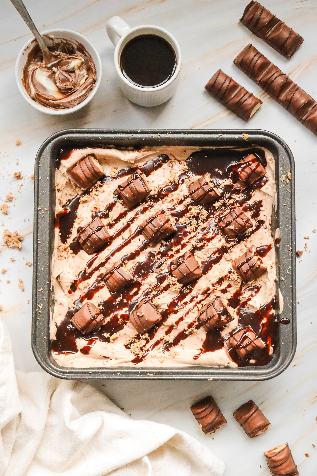 A Kinder Bueno No Bake Cake chocolate dessert in a pan, topped with candy bars and decadent chocolate drizzle, is perfectly presented with coffee and a cup nearby.