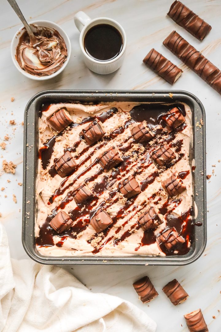 Square pan of chocolate dessert topped with candy bars and chocolate drizzle, served alongside coffee and Nutella—this irresistible Kinder Bueno No Bake Cake is a decadent treat for any sweet tooth.