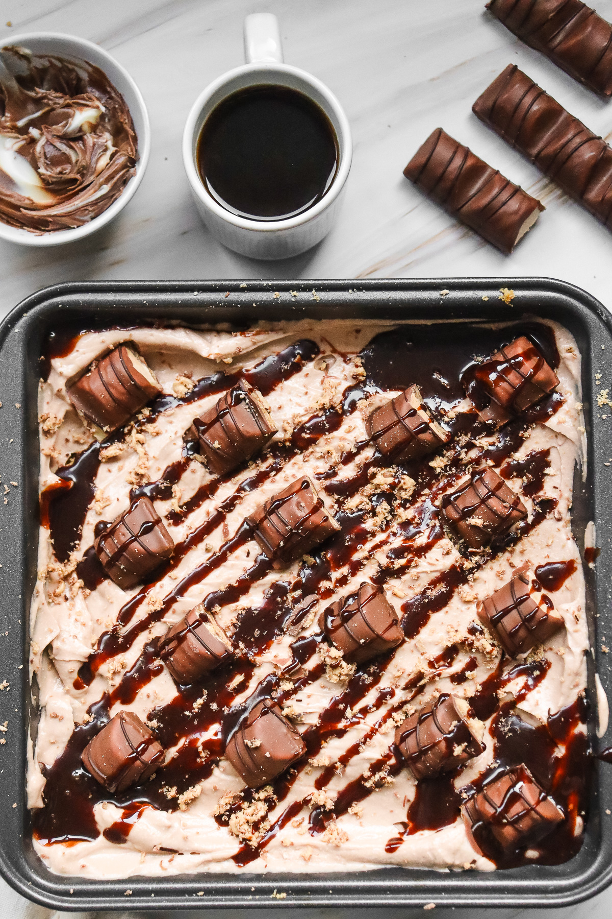 A tray of decadent Kinder Bueno No Bake Cake dessert topped with candy pieces and chocolate drizzle, with coffee and chocolate on the side.