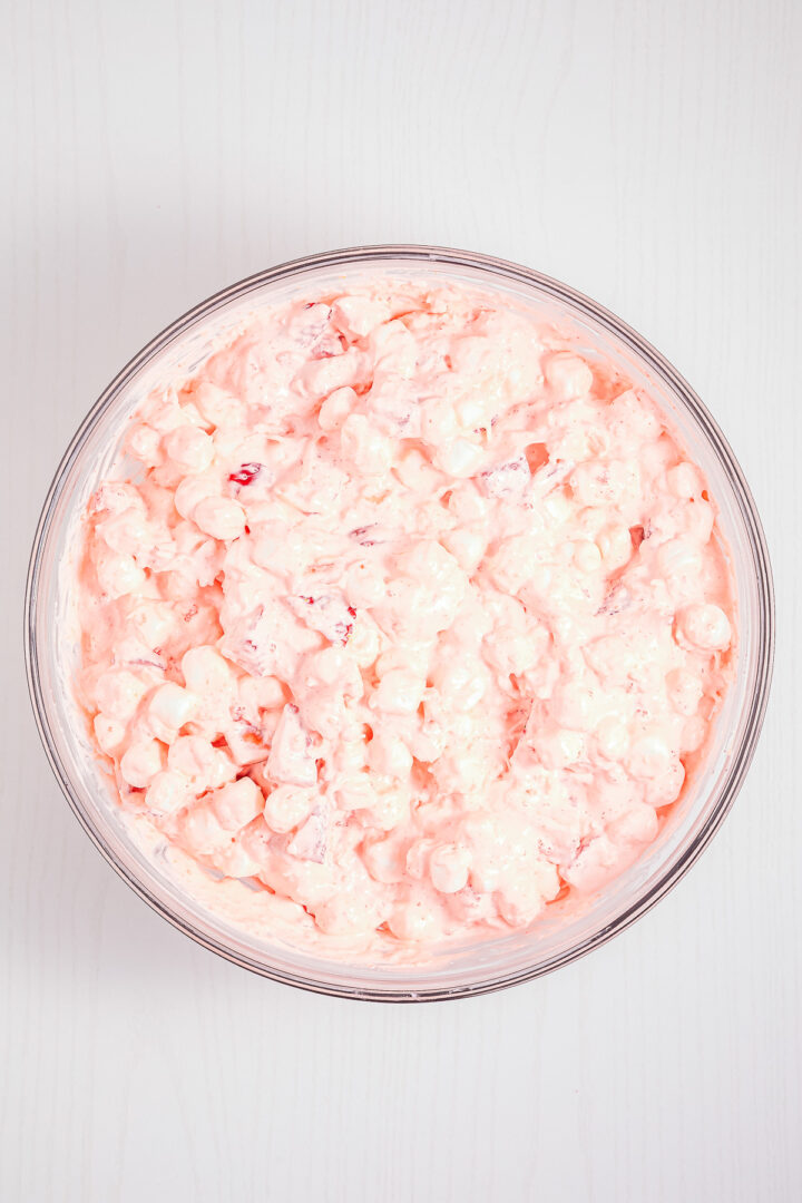 A glass bowl filled with a creamy, pink Strawberry Fluff fruit salad mixture on a white surface.