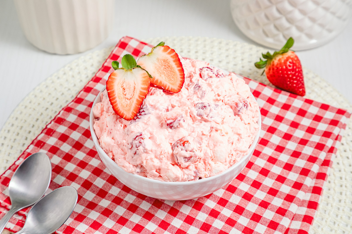 A bowl of Strawberry Fluff salad topped with sliced strawberries sits on a red checkered cloth, with two spoons nearby.