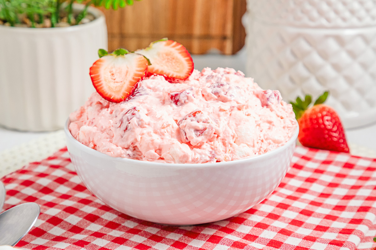 A white bowl of Strawberry Fluff salad topped with sliced strawberries sits on a red checkered cloth.