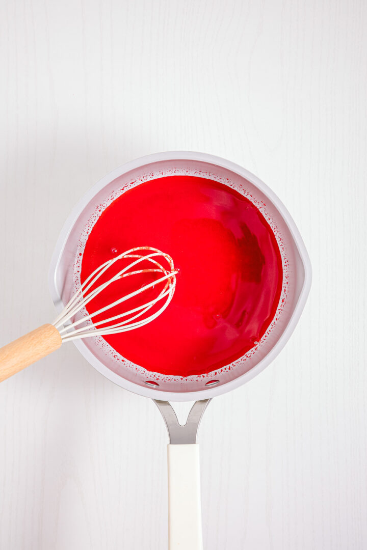A saucepan with red liquid, resembling Strawberry Fluff, and a whisk on a white surface, viewed from above.