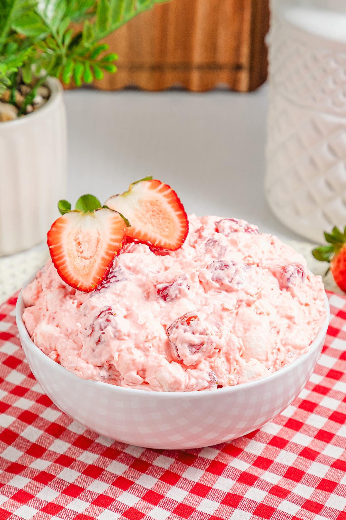 A bowl of pink Strawberry Fluff salad topped with sliced strawberries rests on a red checkered cloth.