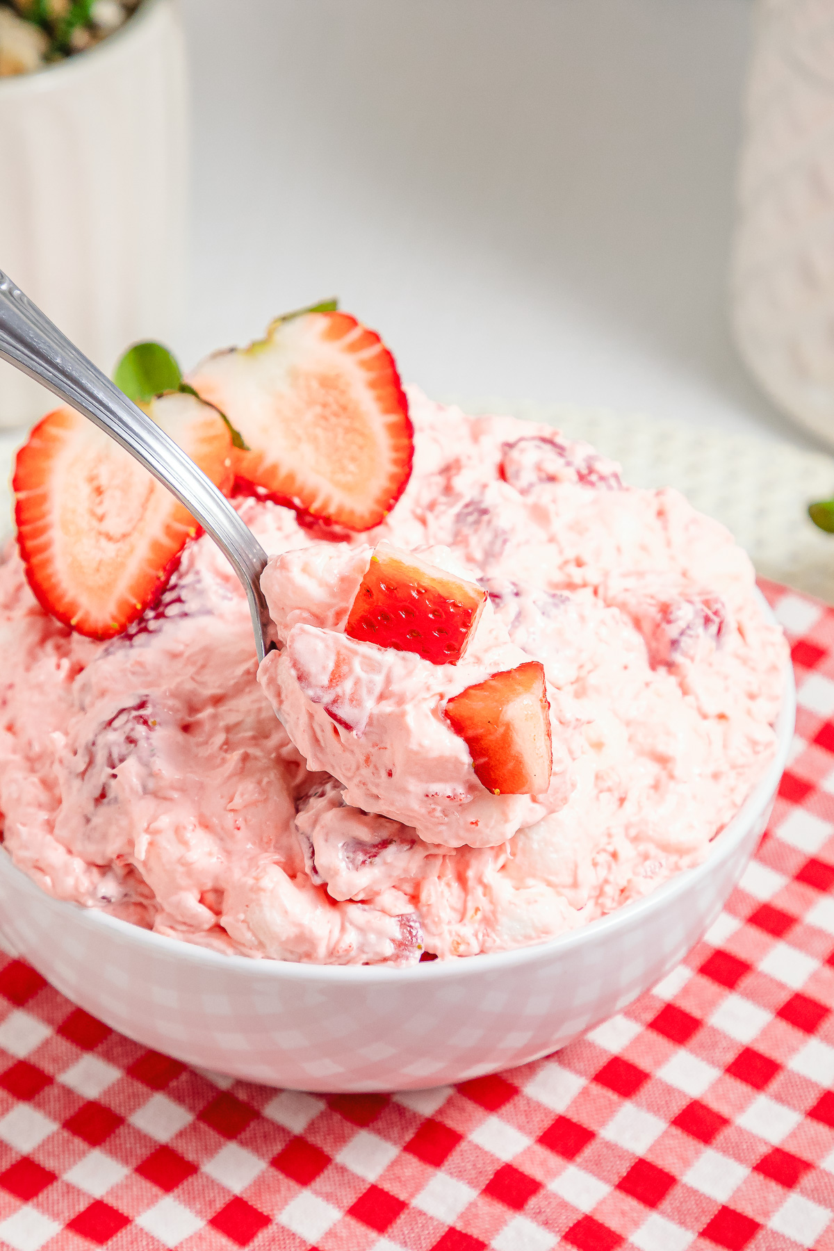A bowl of creamy Strawberry Fluff salad, topped with sliced strawberries, sits invitingly on a red checkered cloth.