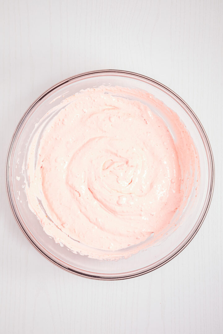 A glass bowl filled with pale pink Strawberry Fluff whipped mixture sits on a white surface.