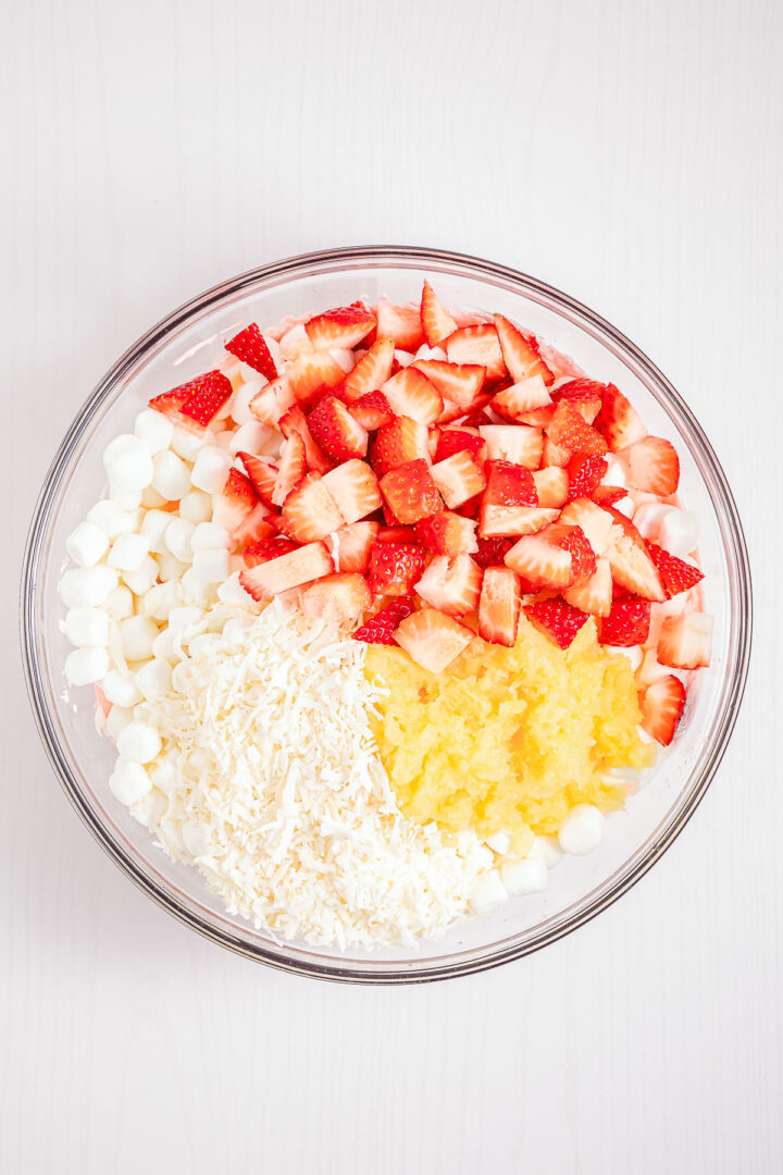 A glass bowl with strawberries, mini marshmallows, shredded coconut, and crushed pineapple on a white table—an irresistible Strawberry Fluff dessert ready to enjoy.