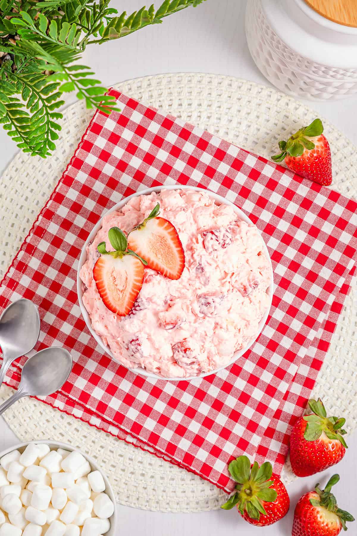A bowl of creamy Strawberry Fluff salad, topped with sliced strawberries, rests on a red checkered napkin.
