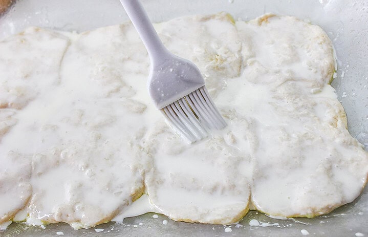 A pastry brush spreading white liquid over uncooked dough pieces in a baking dish, preparing the base for a delectable Strawberry Shortcake Casserole.