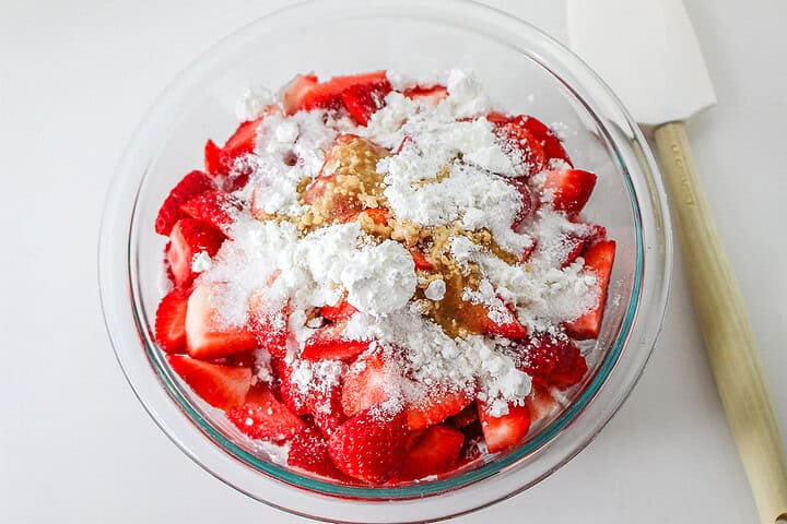 Sliced strawberries in a glass bowl topped with powdered sugar and brown sugar, next to a white spatula—perfect for adding to your favorite Strawberry Shortcake Casserole.