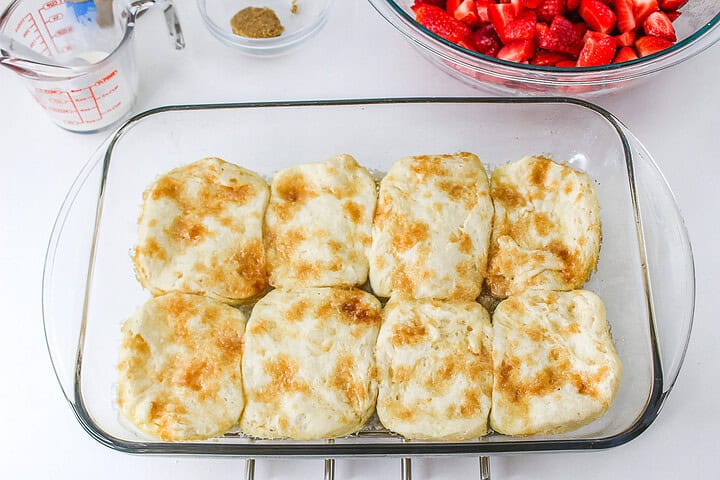 Eight golden-brown biscuits in a glass pan, ready to become a delicious Strawberry Shortcake Casserole, with chopped strawberries and measuring cups nearby.