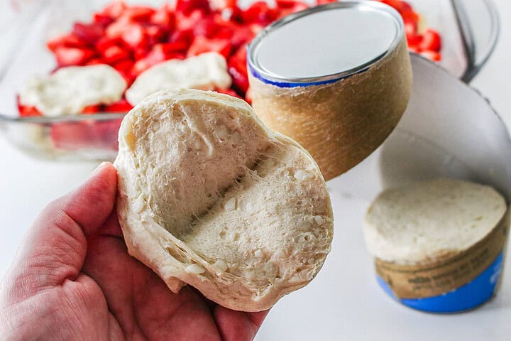 A hand holds unbaked biscuit dough near sliced strawberries and an opened tube of biscuit dough—perfect ingredients for preparing a delicious Strawberry Shortcake Casserole.