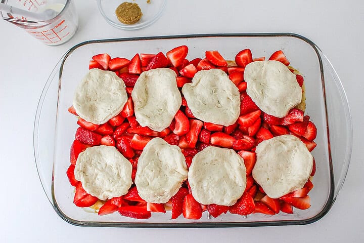 Glass dish with sliced strawberries topped with seven raw biscuit dough rounds, ready to bake into a delicious Strawberry Shortcake Casserole.