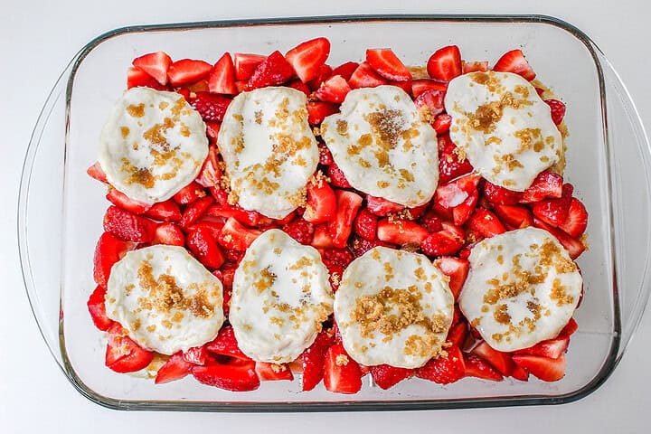 A glass dish with sliced strawberries and biscuit dough, topped with sugar and butter, ready to be baked into a delicious Strawberry Shortcake Casserole.