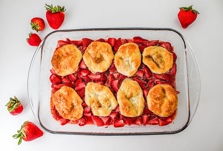 Glass dish with baked biscuits on top of sliced strawberries, creating a delicious Strawberry Shortcake Casserole, surrounded by whole strawberries on a white surface.