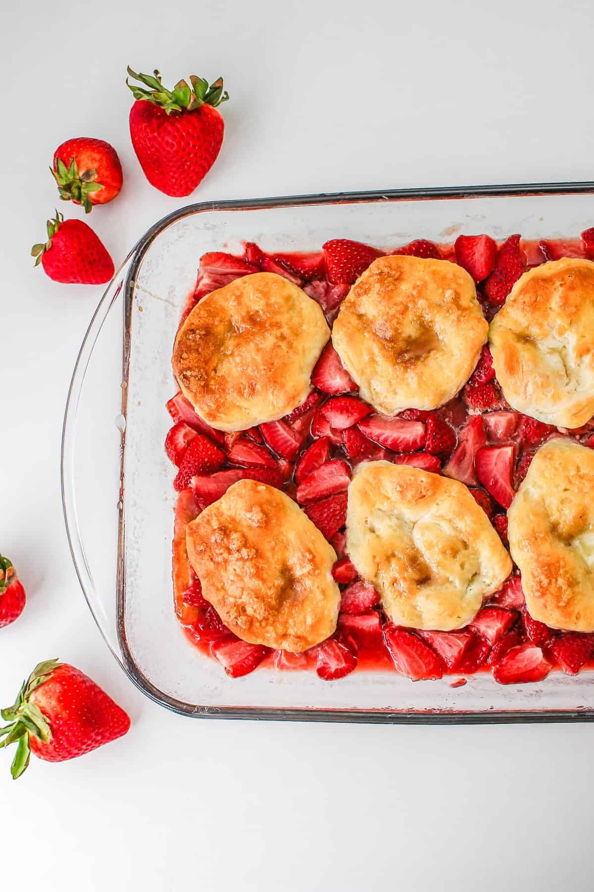 Glass dish with baked biscuits on sliced strawberries, creating a tempting Strawberry Shortcake Casserole, surrounded by fresh strawberries on a white background.