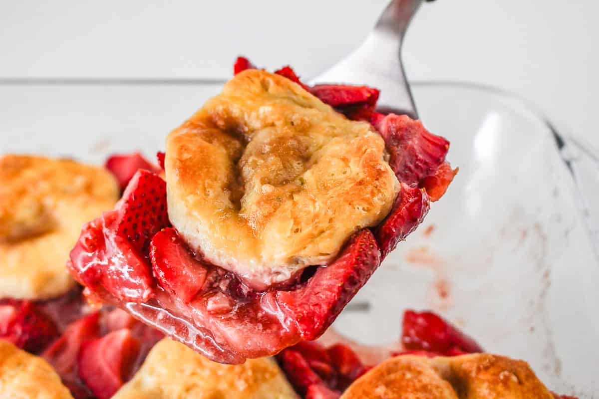 Golden biscuit and juicy strawberries on a spatula, lifted from a glass baking dish of Strawberry Shortcake Casserole.