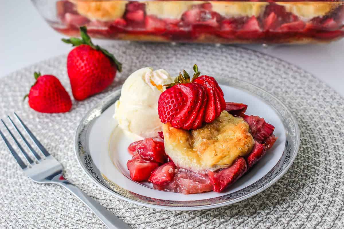 A plate of strawberry cobbler with ice cream, garnished with fresh strawberries, sits beside a fork—evoking the comforting flavors of a classic Strawberry Shortcake Casserole.