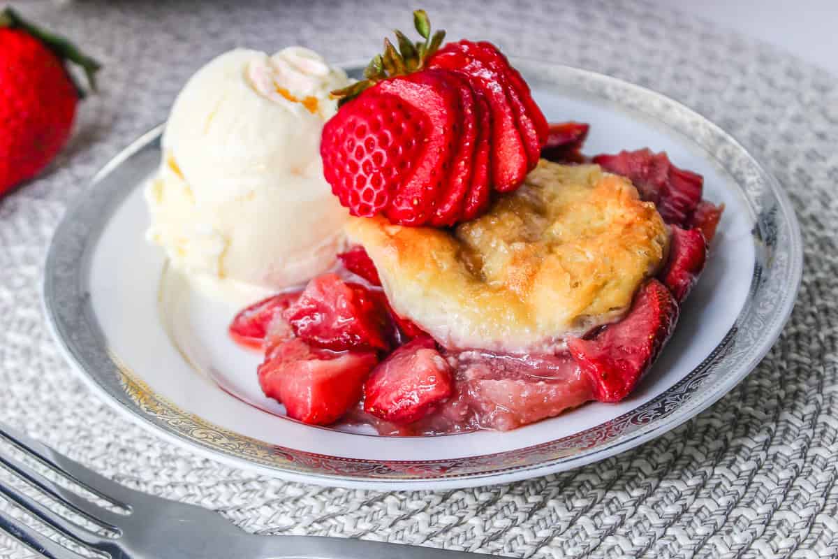 A plate with strawberry cobbler, reminiscent of a Strawberry Shortcake Casserole, topped with a scoop of vanilla ice cream and fresh sliced strawberries.