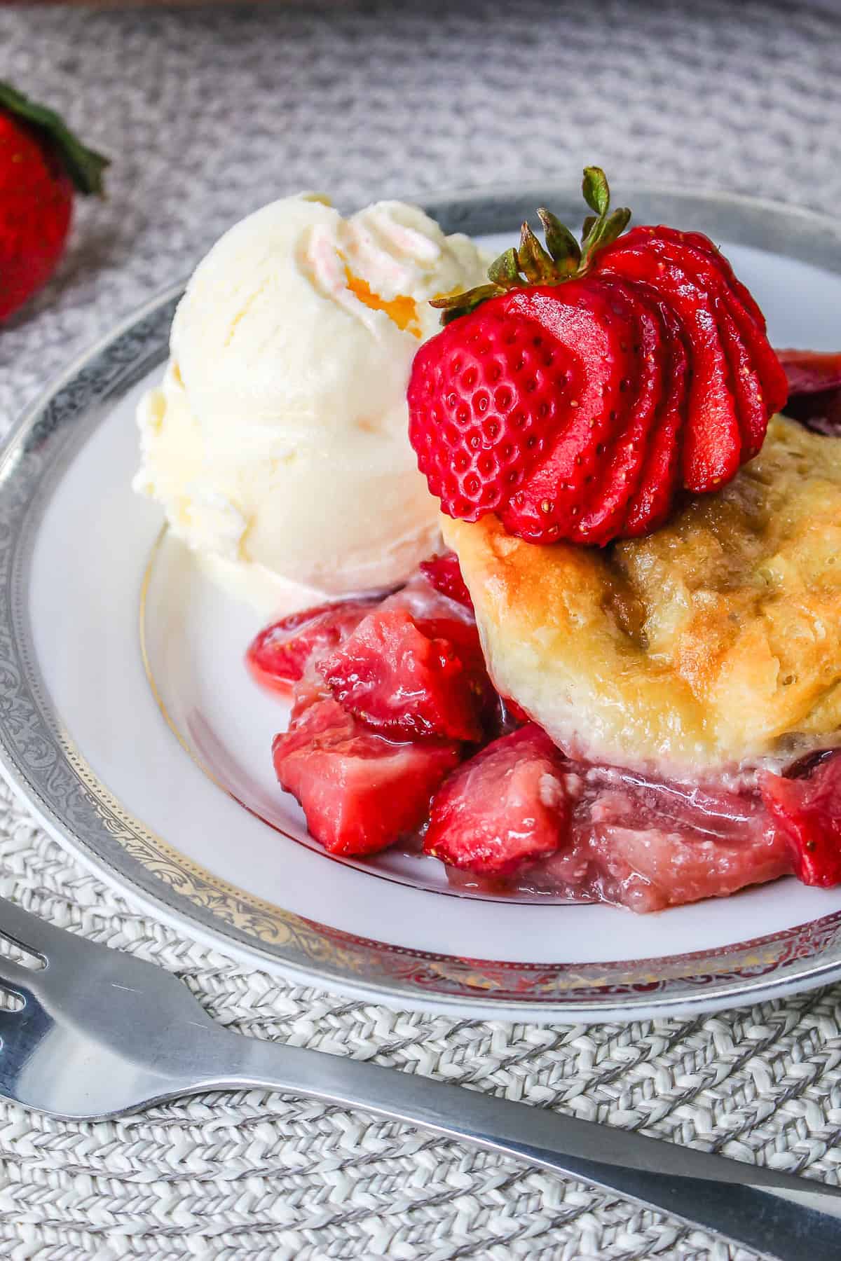 A plate with strawberry cobbler, vanilla ice cream, and a sliced strawberry on top, next to a fork—this delightful presentation evokes all the comfort of a homemade Strawberry Shortcake Casserole.