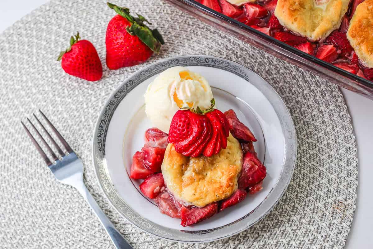A plate with Strawberry Shortcake Casserole, creamy ice cream, and sliced strawberries, with whole strawberries nearby.