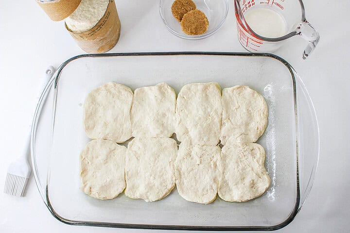 Eight raw biscuit dough rounds are arranged in a glass baking dish, with baking ingredients nearby—ready to be transformed into a delicious Strawberry Shortcake Casserole.