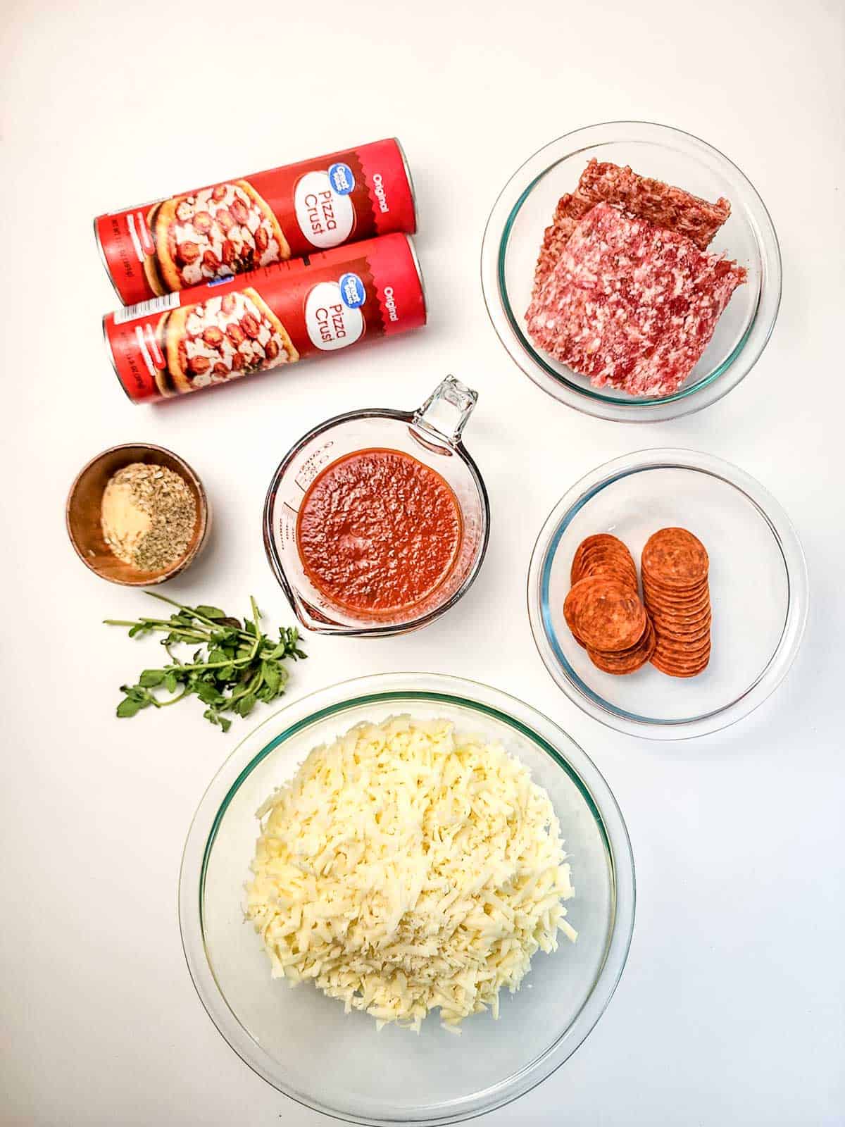Pizza dough, mozzarella, pizza sauce, sausage, pepperoni, breadcrumbs, and herbs on a white background.