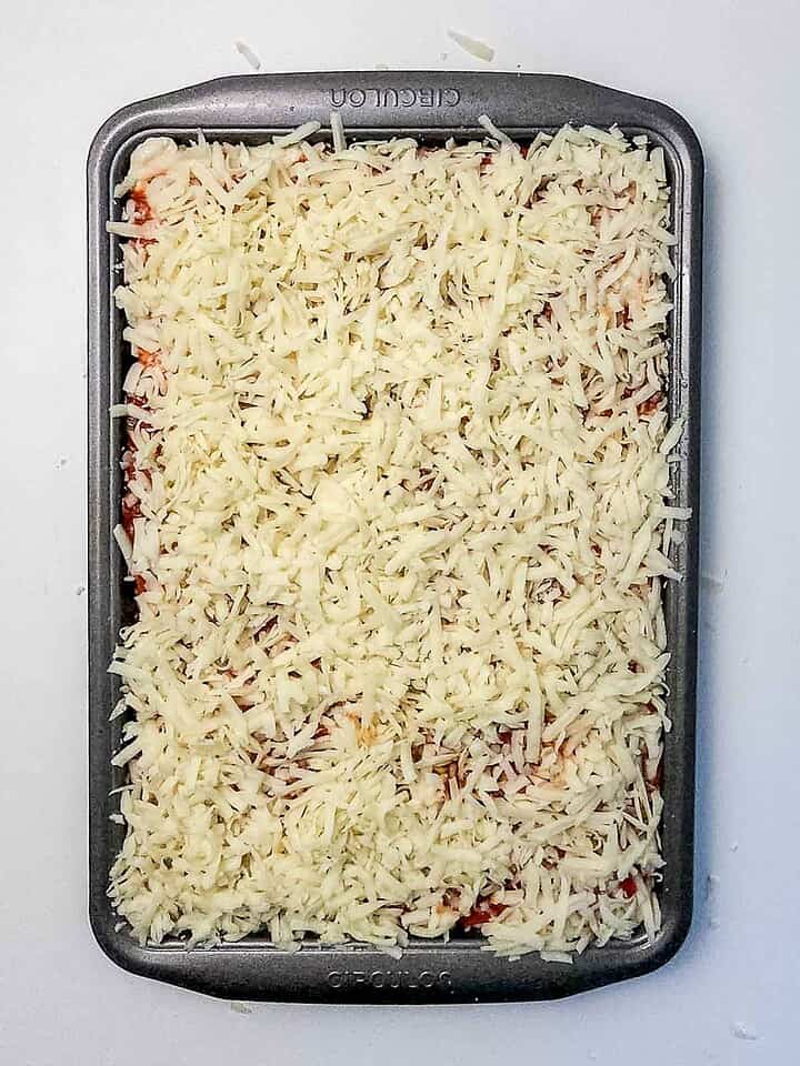 Rectangular baking dish filled with shredded cheese on top of an uncooked Italian Sausage Sheet Pan Pizza casserole.