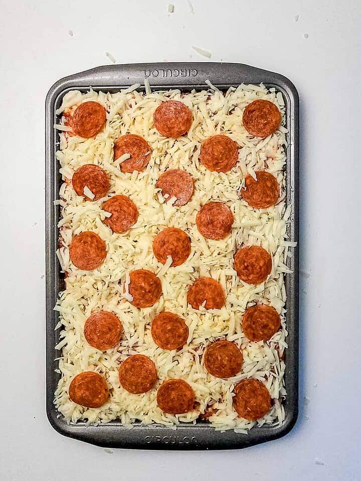 A rectangular baking pan filled with uncooked Italian Sausage Sheet Pan Pizza, generously topped with mozzarella cheese, savory Italian sausage, and pepperoni slices.