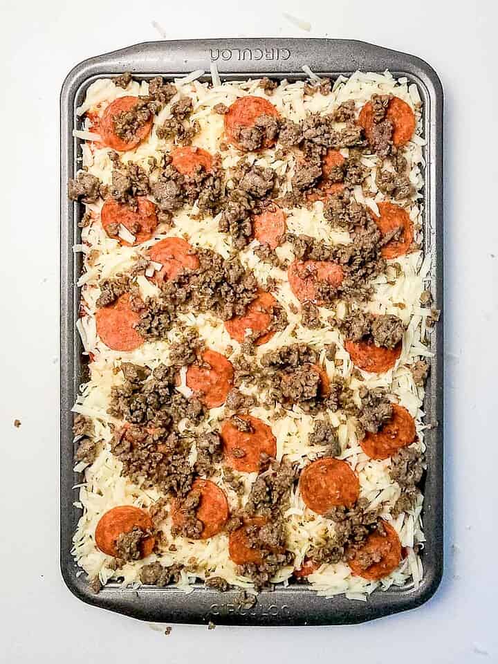 A sheet pan covered with uncooked Italian Sausage Sheet Pan Pizza, generously topped with cheese, pepperoni slices, and crumbled sausage.