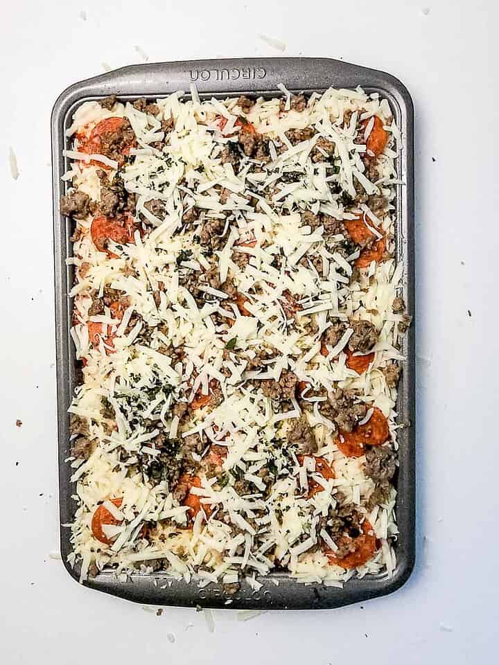 Unbaked Italian Sausage Sheet Pan Pizza in a rectangular pan topped with shredded cheese, savory sausage, pepperoni, and herbs.