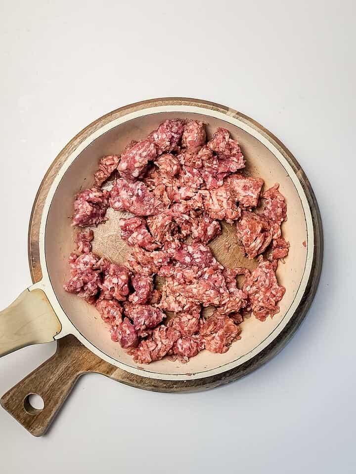 Raw ground meat in a white frying pan with a wooden handle, viewed from above on a white background&mdash;perfect for preparing the Italian Sausage Sheet Pan Pizza you&rsquo;ve been craving.