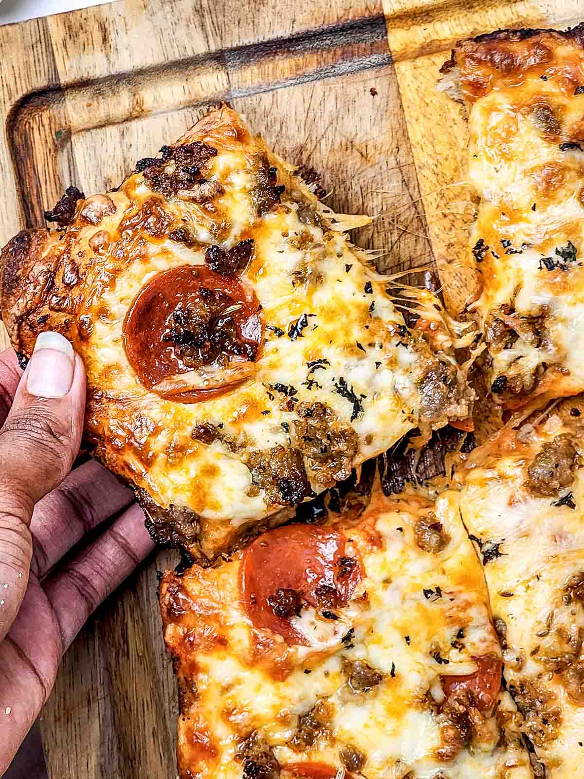 Hand holding a slice of cheesy, square Italian Sausage Sheet Pan Pizza topped with pepperoni and sausage on a wooden cutting board.
