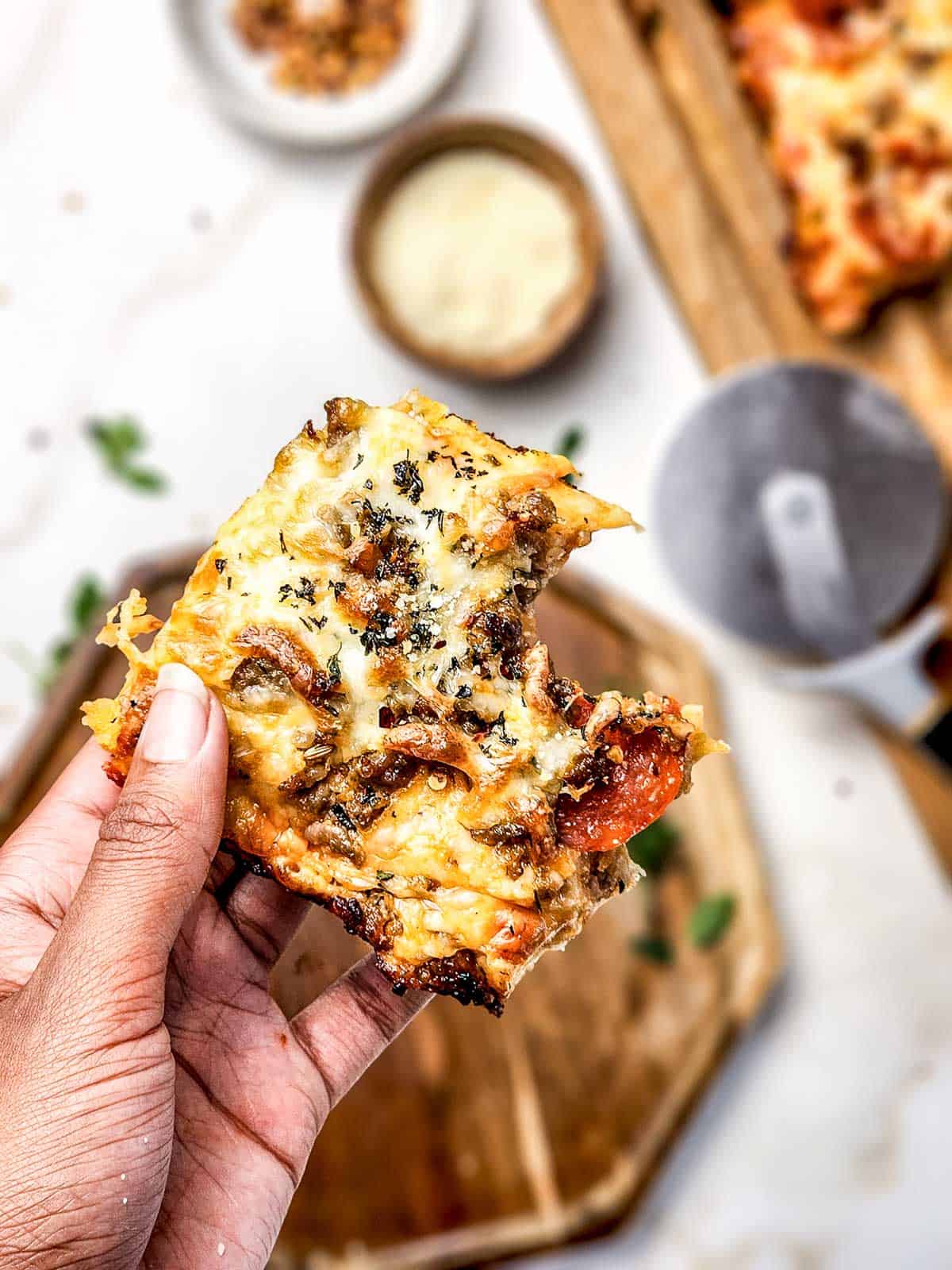 A hand holds a cheesy slice of Italian Sausage Sheet Pan Pizza with a bite taken out, with pizza and savory toppings in the background.