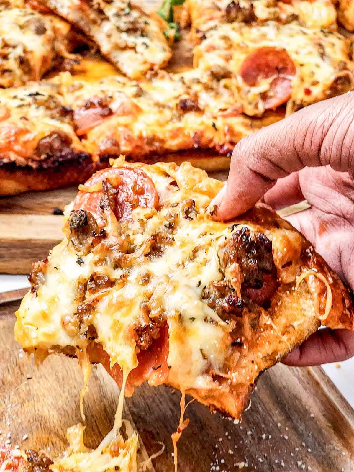 A hand holding a cheesy slice of Italian Sausage Sheet Pan Pizza above a wooden board with more pizza slices.