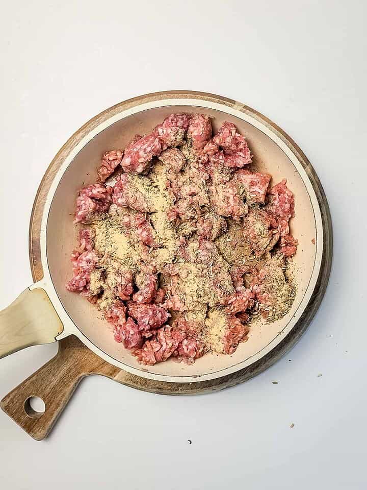 Raw ground meat with seasoning in a beige skillet on a wooden cutting board, viewed from above&mdash;perfectly prepped for making a delicious Italian Sausage Sheet Pan Pizza.