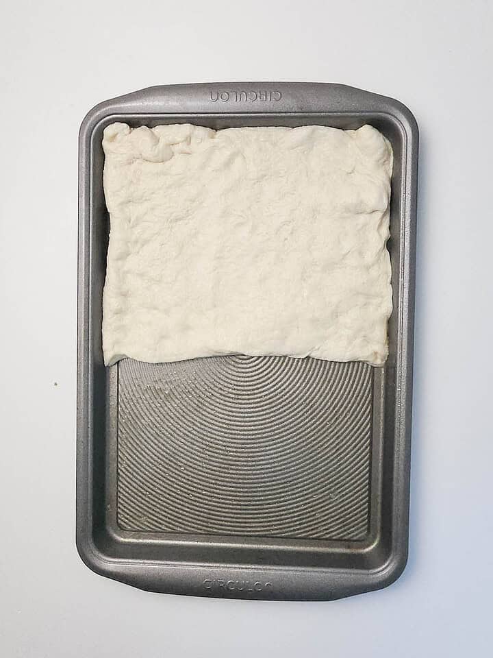 Rectangular baking pan with raw dough stretched to cover only half of the pan, perfect for making Italian Sausage Sheet Pan Pizza, on a white background.