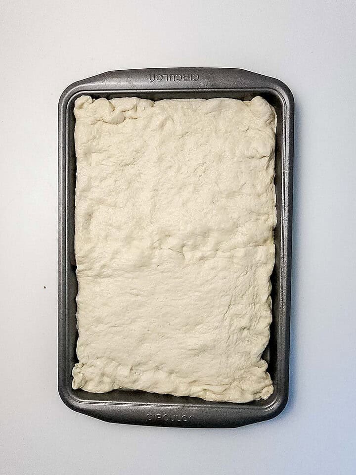 Rectangular baking pan with an even layer of raw dough pressed into the bottom, ready to become a delicious Italian Sausage Sheet Pan Pizza, on a white surface.