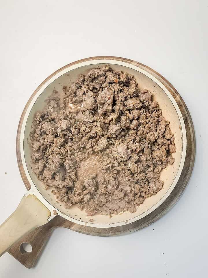 Cooked ground beef in a white skillet with a wooden handle, placed on a round wooden board&mdash;perfect for topping your favorite Italian Sausage Sheet Pan Pizza.