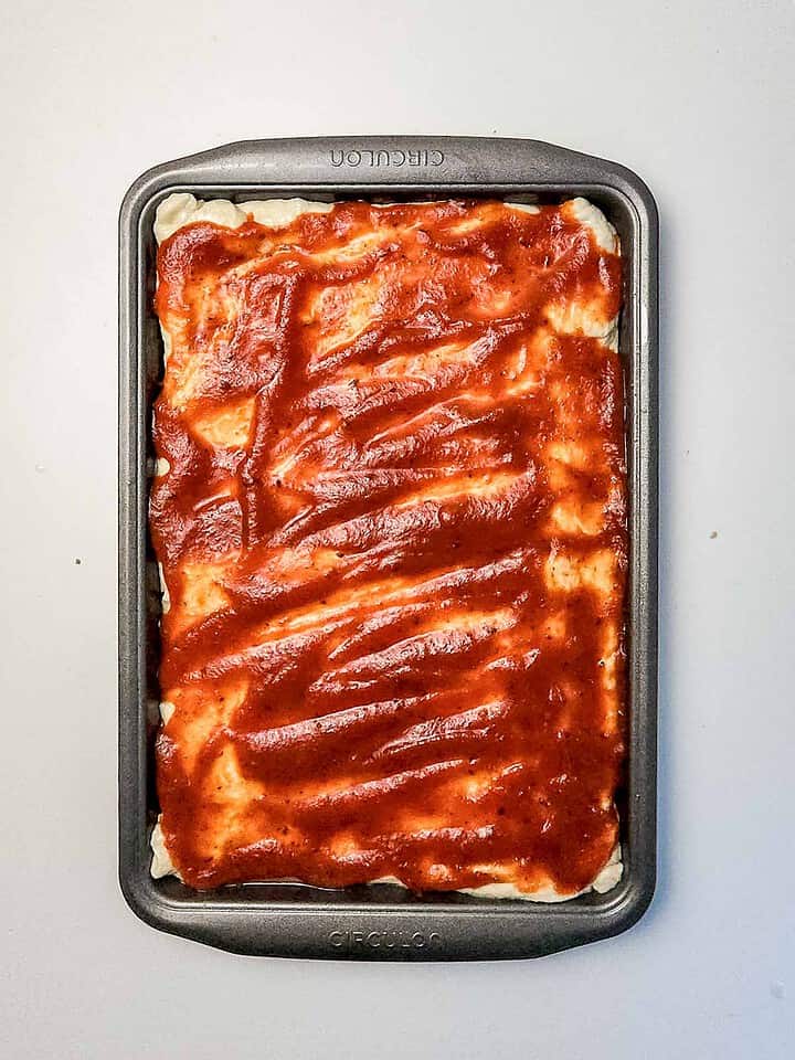 Rectangular baking pan with pizza dough covered in red tomato sauce, ready to be transformed into a delicious Italian Sausage Sheet Pan Pizza, on a plain white surface.