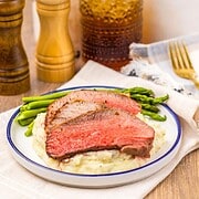 Sliced Air Fryer Eye of Round Roast on mashed potatoes with green beans, beside salt, pepper, and a drink on a wooden table.